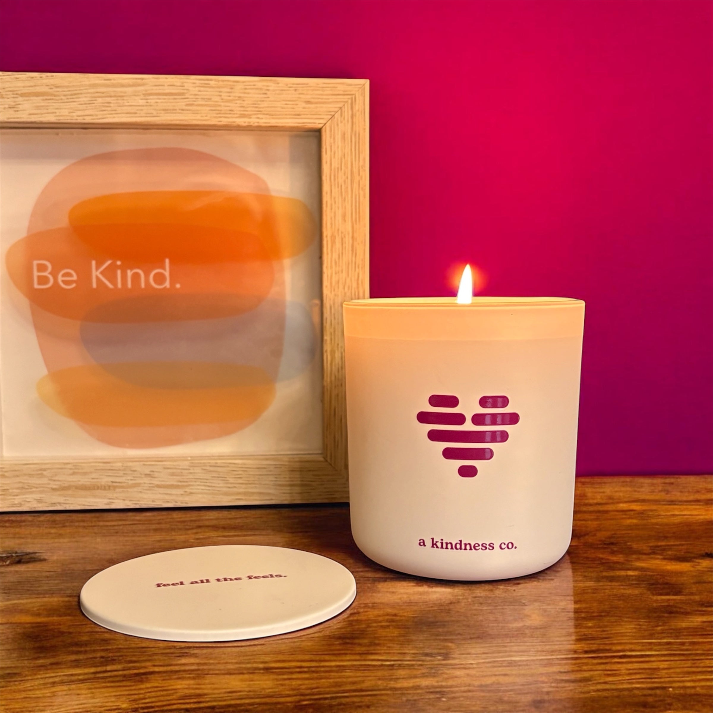 White A Kindness Co candle with heart icon sitting on table with Be Kind sign next to it.